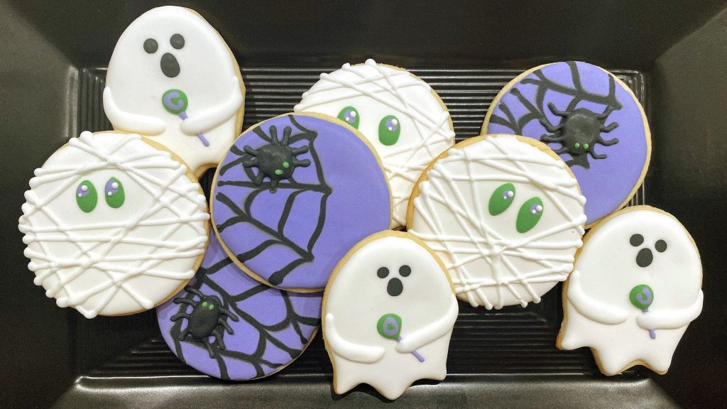 Halloween cookies on a black tray including ghosts, mummies and spiders on a web in purple, green and white.