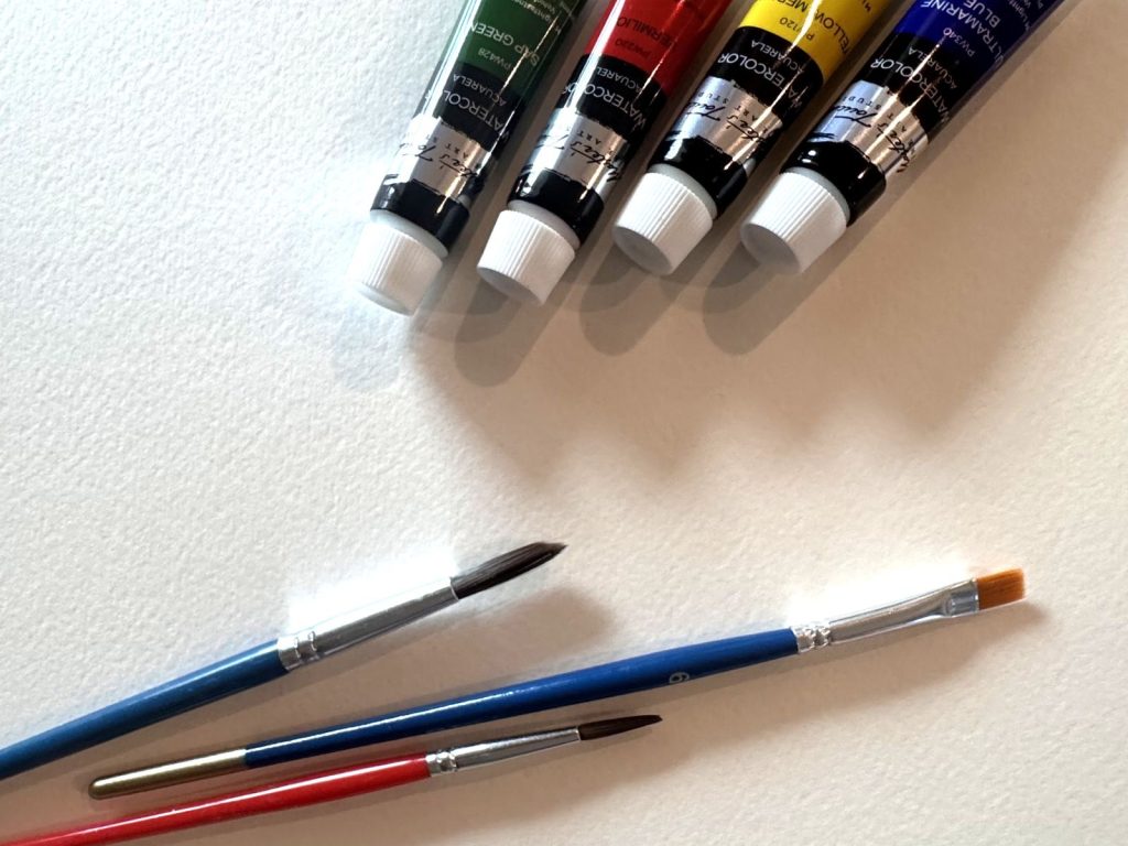 Tubes of watercolor paint and paint brushes sitting on paper.