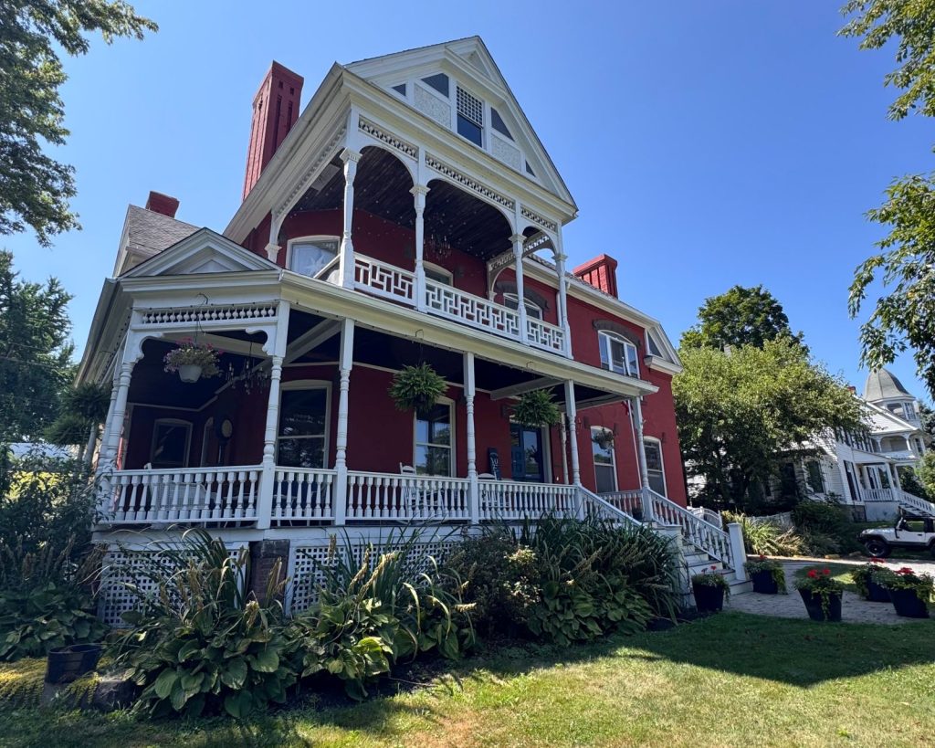Red brick house with two story white porches, and lots of greenery in the yard.