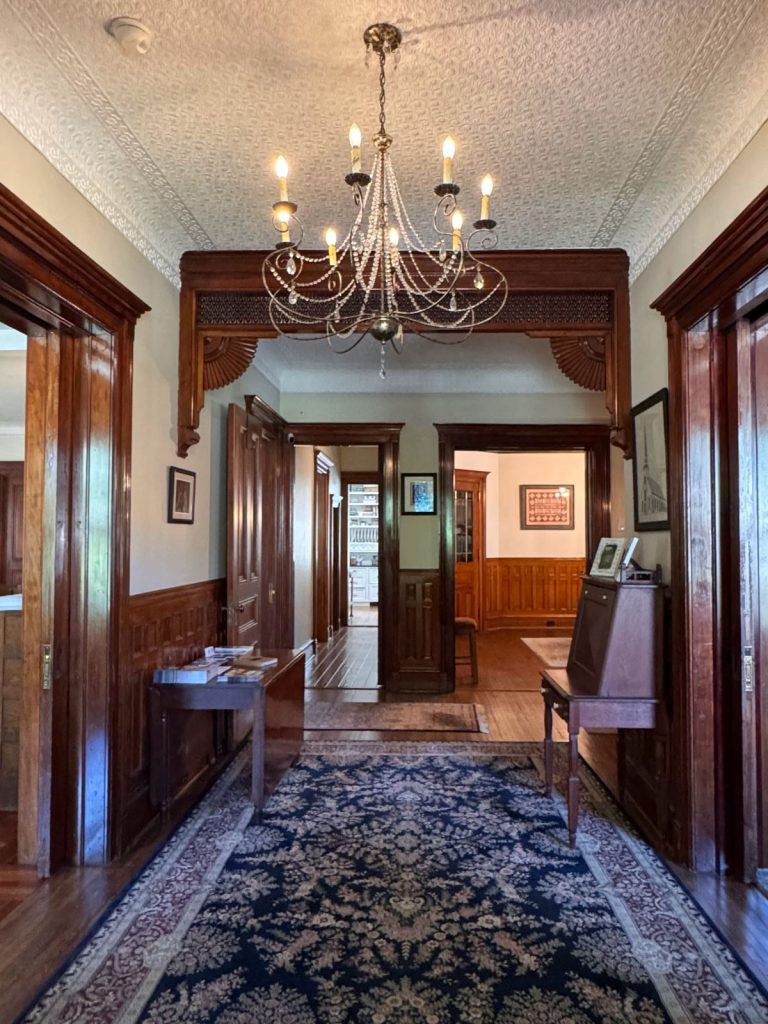 Entry Hall at Hatch House Bed and Breakfast with chandelier, ornate woodwork, detailed ceiling and doorways beyond leading to dining room and kitchen.