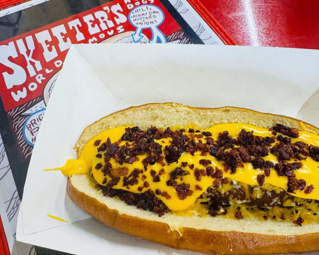 Hot dog with cheese and bacon in a paper bowl with "Skeeter's world famous" menu in background.